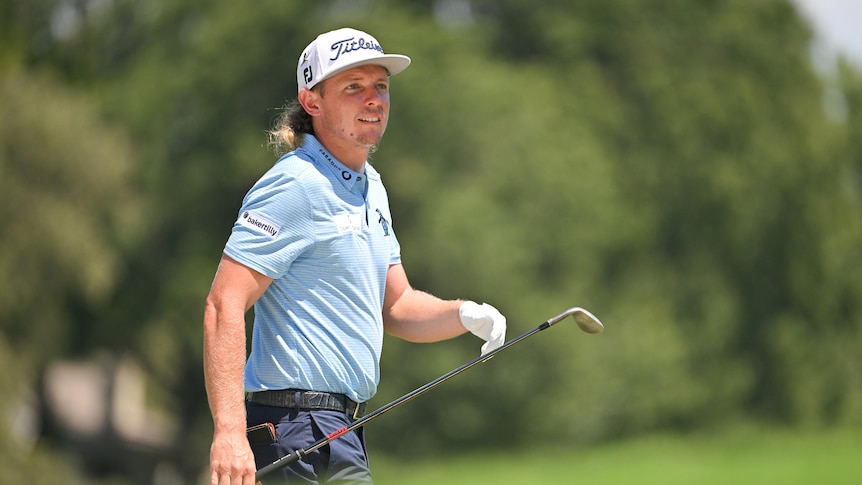 An Australian golfer walks along, holding an iron in one hand as he looks down the fairway towards the green.