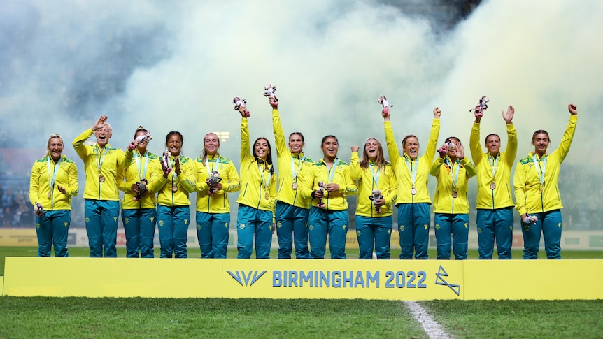 Australia's women's rugby sevens team stand on podium waving with their gold medals as firework smoke drifts behind them.