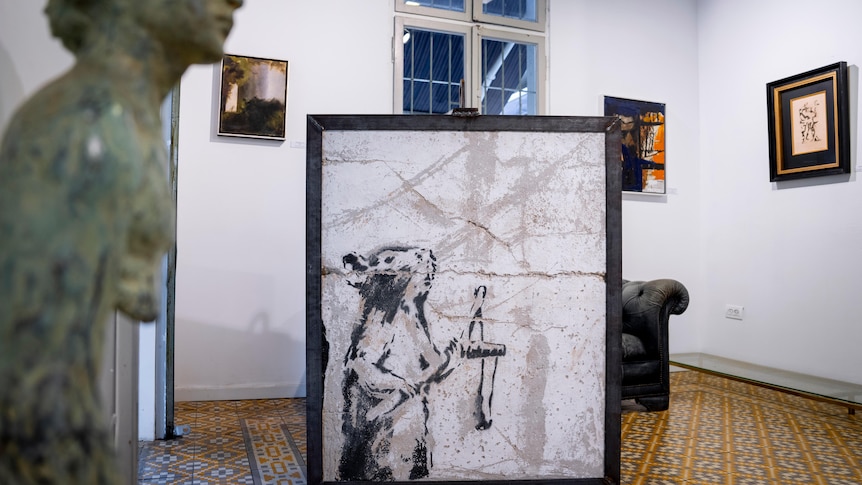 A piece of art featuring a rat sits in a frame on the floor in a room with art on the walls.