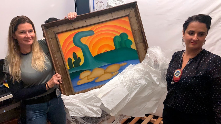 Two women stand either side of Tarsila do Aarel's painting.