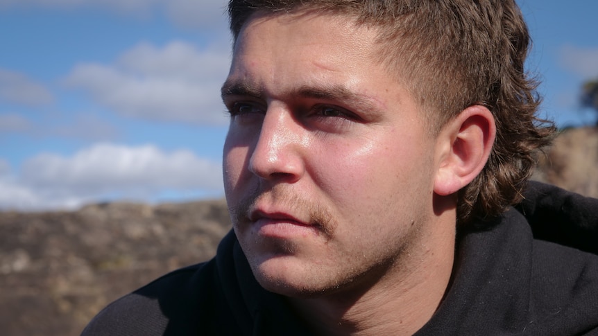 Close up portrait of young man with serious expression with the sky and mountains in the foreground.
