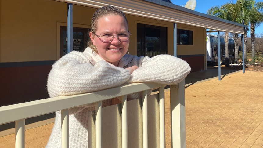 A woman wearing a white jumper leans against a fence in front of a sign that says Rivergum Christian College.