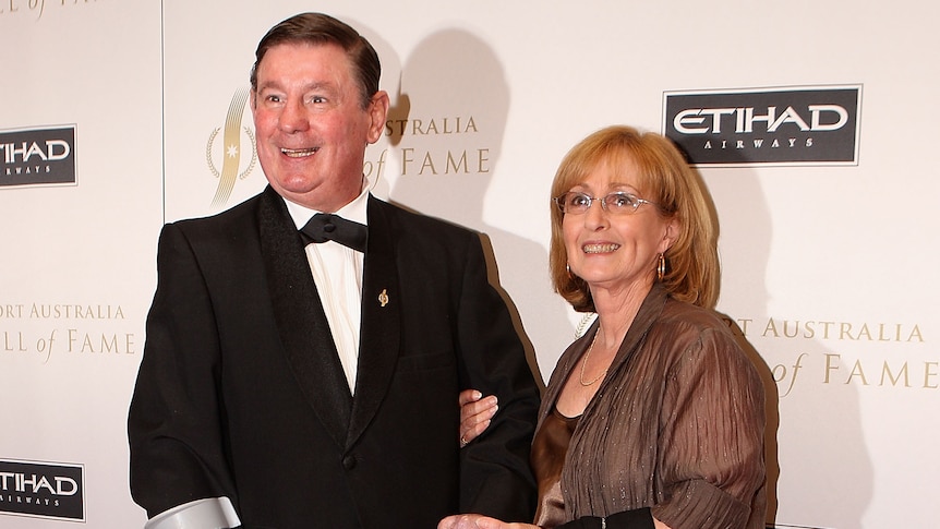 A man wearing a tuxedo smiles for the camera, using a crutch as he stands with a woman before a sporting ceremonial dinner.