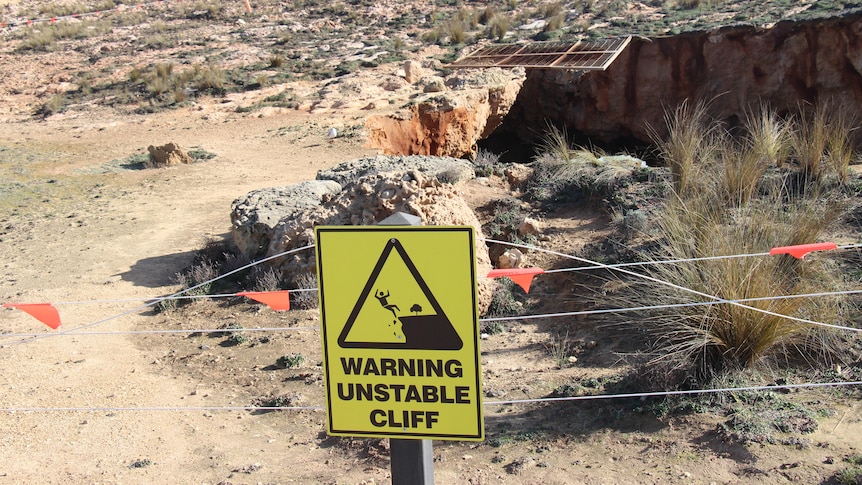 A yellow sign reading 'warning unstable cliff' with a barricade and coastal hole behind