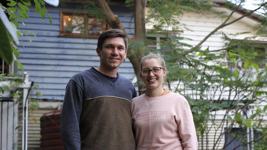 Man and woman smiling in front of their weatherboard home painted blue and cream, near a tree.
