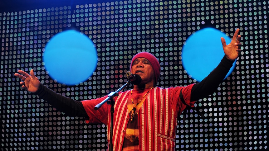 Archie Roach sings on stage with his arms outstretched and his eyes closed.