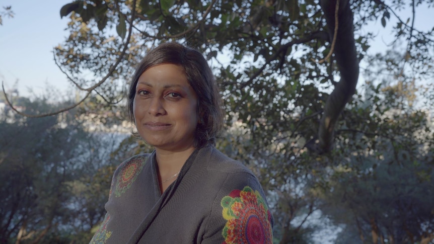 Indira Naidoo has short hair, half smile, wears a grey knit with colourful print, stands under a giant tree.