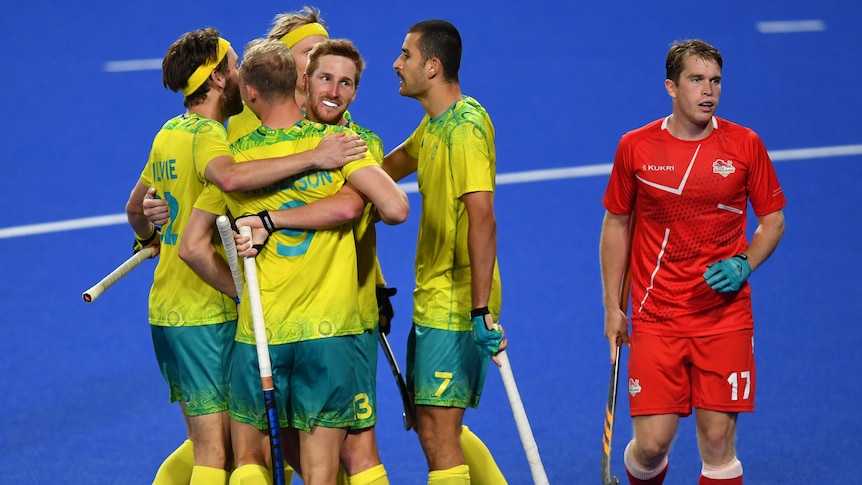 A group of Australian men's hockey players hug after a goal, as a dejected England player stands by.