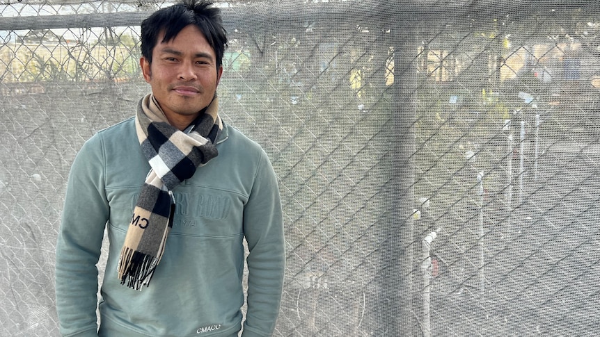 A man in a green jumper standing in front of a chain link fence.