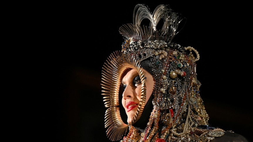 A man wearing a heavy-looking piece of headwear, draped with jewels, stares out of the darkness to the side of the frame..