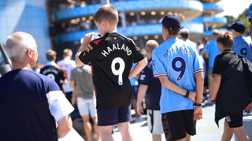 Two young Manchester City fans, one drinking from a bottle, in Erling Haaland jerseys with other fans outside the stadium