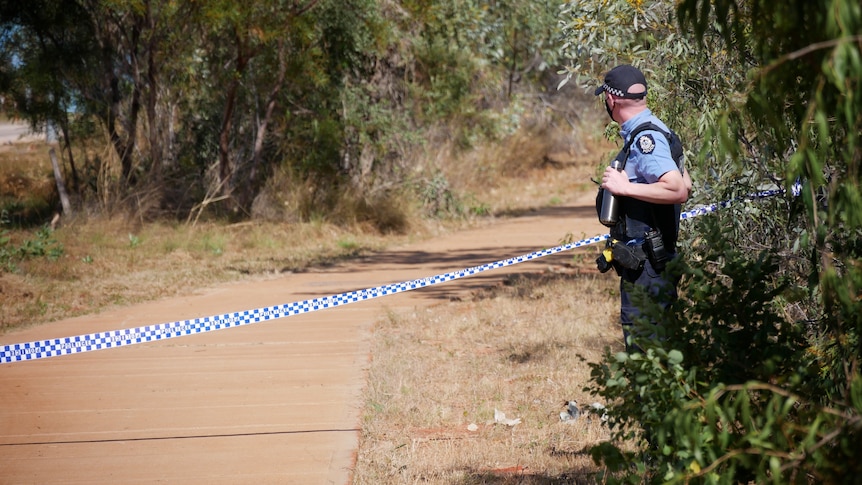 A uniformed officer with police tape stretched across a path with bushes.
