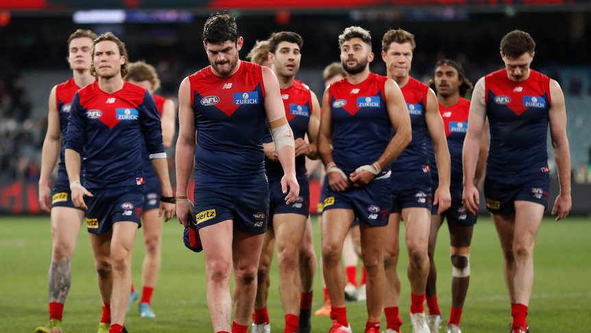 A group of Melbourne AFL players walk off the ground looking disappointed after a match at the MCG.