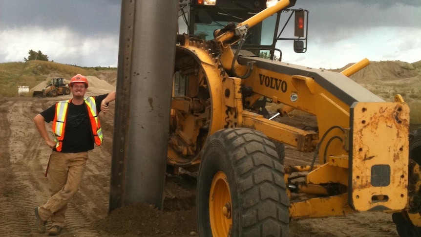 A smiling man in high-vis leans against a huge girder sticking out of the earth near a digger.