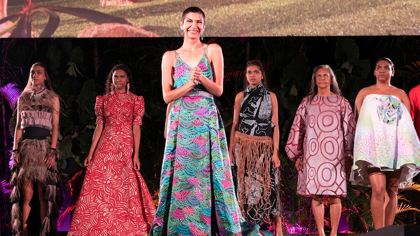 A group of fashion models stand in a line on the stage. A woman in a colourful dress stands in front of them.