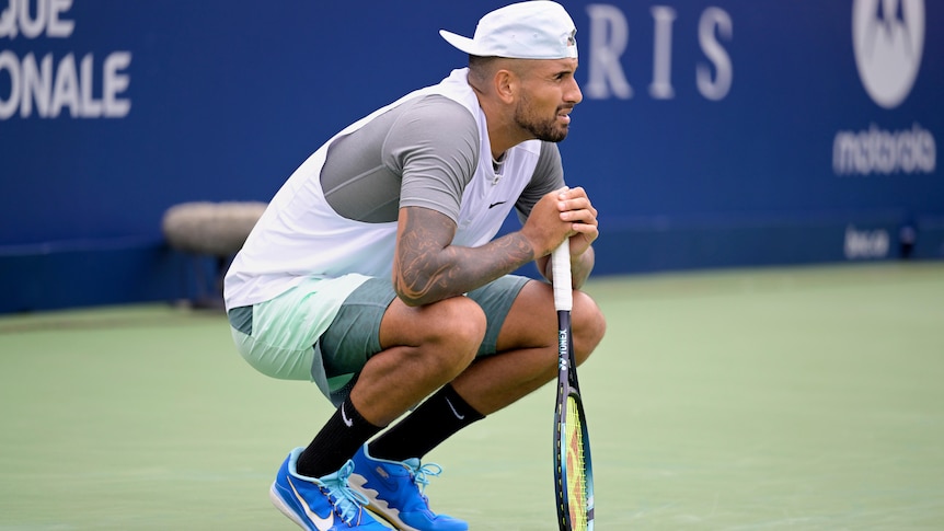 Nick Kyrgios crouches down and leans on his racquet on court