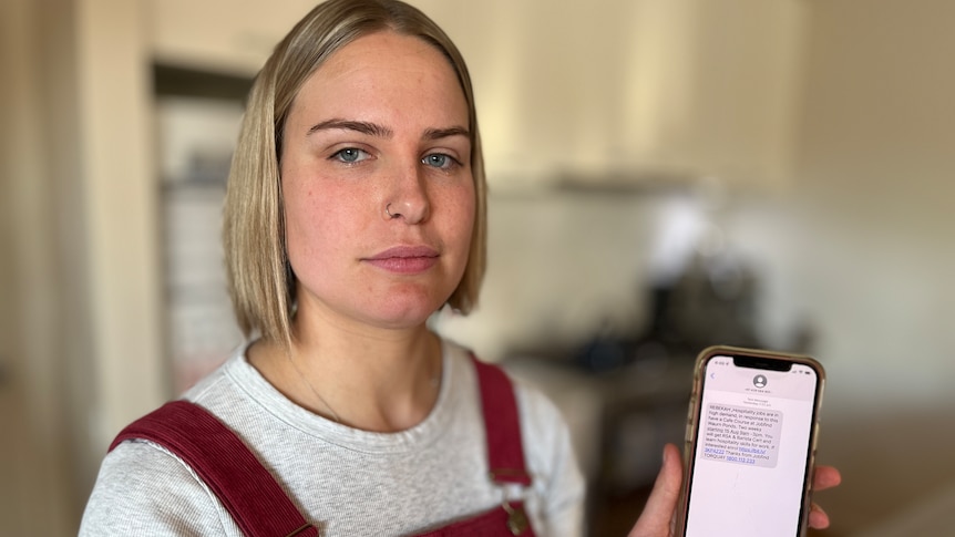 A woman with blonde hair holding up her phone, looking at the camera