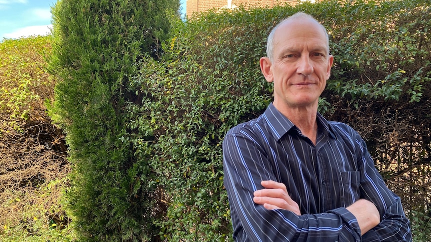 A middle aged man with his arms crossed looking at the camera while standing in front of a bush