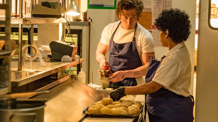 A man acting as a chef is shown holding cooking tools and looking at a woman facing away in a kitchen